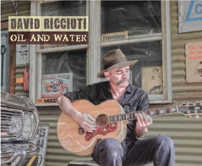 Man plays guitar in front of rustic building.